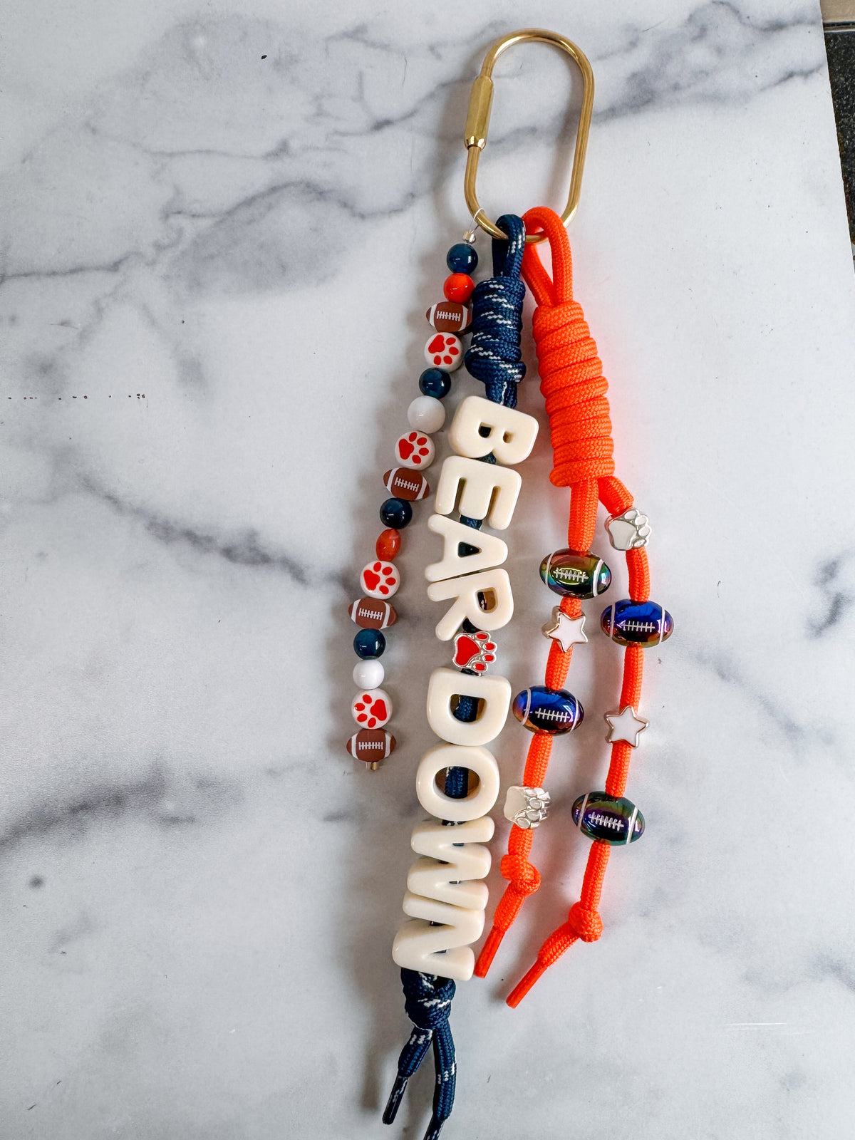 A custom carabiner bag charm keychain with a gold carabiner clip, featuring white letters 'BEAR DOWN' text charm, orange and navy blue cords, and decorative orange, white, and blue beads.