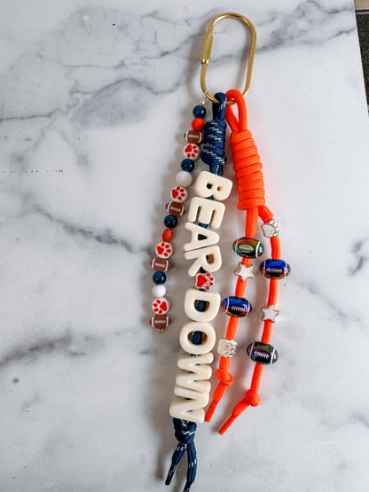 A custom carabiner bag charm keychain with a gold carabiner clip, featuring white letters 'BEAR DOWN' text charm, orange and navy blue cords, and decorative orange, white, and blue beads.