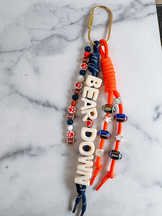 A custom carabiner bag charm keychain with a gold carabiner clip, featuring white letters 'BEAR DOWN' text charm, orange and navy blue cords, and decorative orange, white, and blue beads.